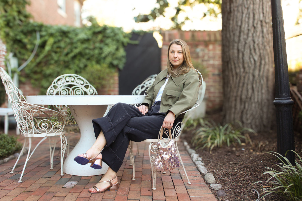 woman sitting in chair wearing a green Ganni Jacket, Black Tibi jeans, and rose gold metallic Marion Parke sandals